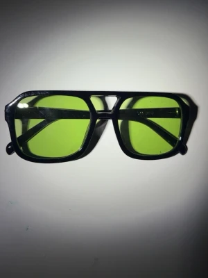 Svarta solglasögon med gröna glas - Snygga solglasögon med svart, kantig båge och ljusgröna glas. Glasögonen har en modern och trendig design som sticker ut. Perfekta för dig som vill ha en cool accessoar till sommaren.