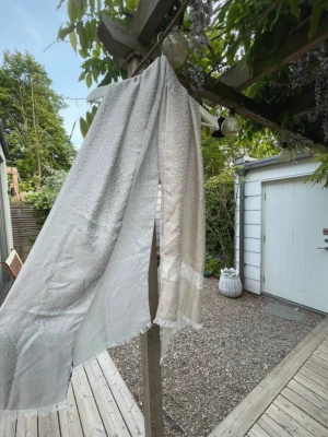 Ljusbeige/ grå  GUESS sjal - Säljer en elegant ljusbeige/ grå sjal med diskret mönster i vitt och grått. Sjalen är tunn och har en lätt glansig yta, vilket ger ett exklusivt intryck. Perfekt att svepa runt halsen för en stilren look. Äkta från guess! 