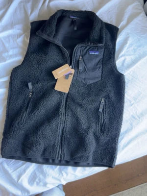 Patagonia väst - Säljer en svart fleeceväst från Patagonia med dragkedja framtill, två sidofickor med dragkedja och en bröstficka med logga. Västen har hög krage och mjukt, fluffigt material som ger en chill look. Perfekt att slänga över en hoodie eller t-shirt.