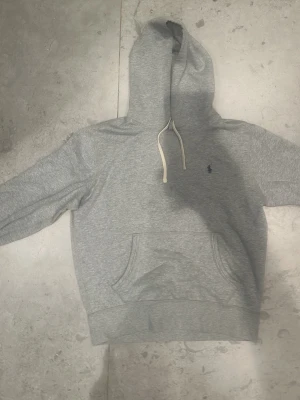 Grå hoodie från Polo Ralph Lauren - Klassisk grå hoodie från Polo Ralph Lauren med huva och vita dragsnören. Har en känguruficka framtill och den ikoniska lilla mörkblå loggan broderad på bröstet. Tillverkad i mjuk bomullsblandning som känns skön mot huden. Perfekt för en avslappnad och stilren look.