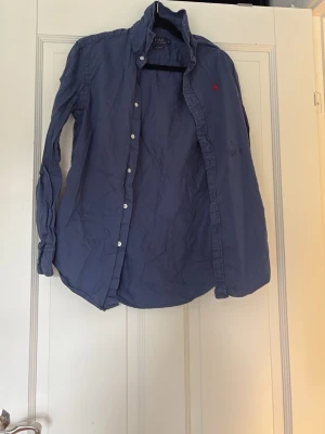 Blå skjorta från Polo Ralph Lauren ca 12 år  - Klassisk blå skjorta från Polo Ralph Lauren med relaxed fit och vit knappstängning. Skjortan har lång ärm och den ikoniska röda logotypen broderad på bröstet. Perfekt för en stilren och avslappnad look.