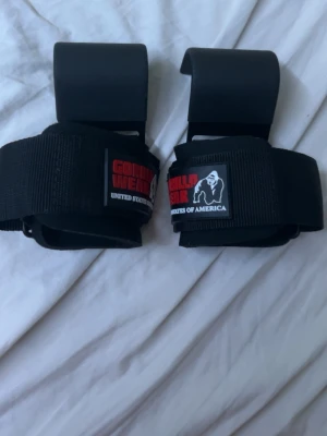 Svarta lyftkrokar Gorilla Wear - Säljer ett par svarta lyftkrokar från Gorilla Wear. De har breda kardborreband och metallkrokar för extra grepp vid styrketräning. Perfekta för tunga lyft och gympass. Märket syns tydligt på banden med röd och vit logga. Helt nya aldrig använda bara testade 