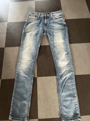 Ljusblå slim Nudie jeans med slitningar - Säljer ett par ljusblå slim fit jeans med snygga slitningar och tvättade detaljer. Klassisk femficksmodell med bälteshällor och knappgylf. Jeansen är tillverkade i denim och har en modern, smal passform som sitter snyggt.