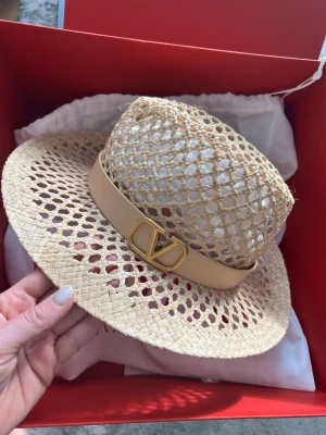 Valentino stråhatt med guldlogo - Snygg stråhatt från Valentino med bred, flätad brätte och öppen, nätliknande krona. Beige läderband med guldfärgad V-logga framtill ger en lyxig touch. Perfekt accessoar för soliga dagar och trendiga outfits.