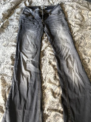 Grå bootcut jeans med slitningar - Säljer ett par grå bootcut jeans med snygga slitningar och klassiska fem fickor. Jeansen har dragkedja och knapp framtill samt bälteshällor. Perfekta för dig som gillar en avslappnad och trendig stil.