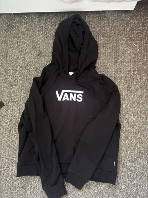 Svart hoodie från Vans med logga - Svart hoodie från Vans med stor vit logga på bröstet. Klassisk modell med huva och ribbade muddar vid ärmslut och nederkant. Tillverkad i mjukt bomullsmaterial som är skönt att ha på sig. Perfekt för en avslappnad streetwear-look.