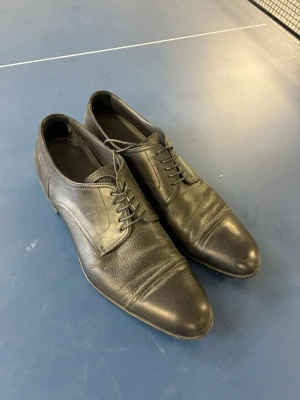 Svarta loafers i läder - Säljer ett par klassiska svarta loafers i äkta läder med rundad tå och diskreta sömdetaljer över tån. Skorna har låg klack och slät sula, perfekta för en stilren look. Passar dig som gillar tidlös och enkel design.