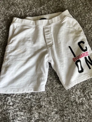 Vita ICON shorts från Dsquared2 - Snygga vita shorts från Dsquared2 med trycket 'ICON' och Dsquared2-loggan i svart och rött på ena benet. Shortsen har elastisk midja, fickor fram och en bakficka. Perfekta för en avslappnad och trendig look.