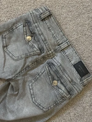 Grå bootcut jeans från Replay - Snygga grå bootcut jeans från Replay med slitna detaljer och markerade sömmar. Jeansen har två bakfickor med lock och silvriga  knappar. Perfekta för dig som gillar en avslappnad men trendig look.