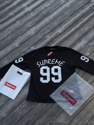 Supreme svart långärmad topp - Supreme långärmad svart topp med stora vita siffror '99' och Supreme-tryck på bröstet samt siffror på ärmarna. Sportig streetwear-look, perfekt för dig som gillar hypebeast-stil. Kommer med originalpåse och plastförpackning.