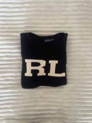 Svart stickad tröja Polo Ralph Lauren - Svart stickad tröja från Polo Ralph Lauren med stora vita RL-bokstäver på bröstet. Tröjan har rund halsringning och ribbstickade muddar vid ärmslut och nederkant. Perfekt för dig som gillar klassisk och stilren design.