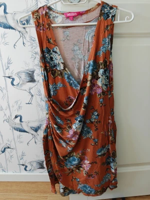 Blommig omlottklänning från Indiska - Säljer en kort blommig omlottklänning från Indiska i varma toner av orange, blått och lila. Klänningen har djup v-ringning, draperad front och är ärmlös. Perfekt för sommarens alla tillfällen!