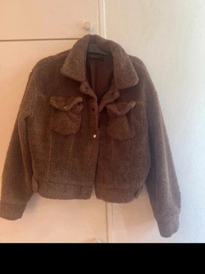 Brun teddyjacka med fickor - Mysig brun teddyjacka med två stora bröstfickor och klassisk krage. Jackan har knappar framtill och ribbade muddar vid ärmslut och nederkant. Fodrad insida för extra komfort. Perfekt för kyliga dagar och ger en trendig look.
