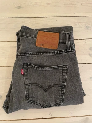 Grå Levi's 501 raka jeans - Säljer ett par klassiska grå Levi's 501 jeans med raka ben och femficksmodell. Jeansen har den ikoniska läderlappen bak i midjan och röd Levi's-tag på bakfickan. Tillverkade i slitstark denim, perfekta för en avslappnad och tidlös stil. Lite sliten längst ned. 