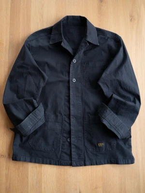 Svart ripstop overshirt, size S - Säljer en svart overshirt  med klassisk krage och tre fickor framtill. Overshirten är tillverkad i slitstarkt bomullstyg och har knäppning med knappar. Perfekt för lager-på-lager och en cool streetstil.