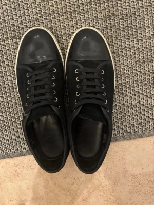 Lanvin svarta sneakers med vit sula - Säljer ett par svarta Lanvin sneakers med vit sula och svart tå i blankt material. Skorna har svarta skosnören och metallöljetter. Klassisk låg modell med stilren design och exklusiv känsla. Dustbags och originalkartong ej medföljer. Lanvins strl:42 men passar 43. Mycket bra skick