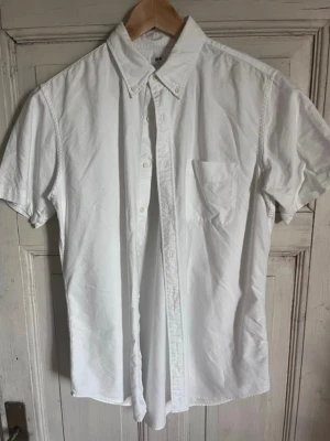 Uniqlo - Vit kortärmad skjorta med knappar - Säljer en klassisk vit skjorta från Uniqlo med korta ärmar och knappar hela vägen fram. Skjortan har en bröstficka och en stilren button-down krage. Perfekt för dig som vill ha en enkel och snygg look.