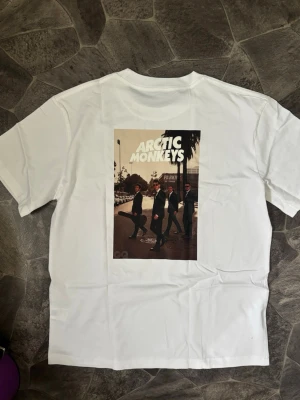 Vit Arctic Monkeys t-shirt - Säljer en vit t-shirt med ett stort tryck på ryggen föreställande Arctic Monkeys i kostym. T-shirten har klassisk passform och rund hals. Perfekt för dig som gillar bandmerch och vill sticka ut med en snygg grafisk design.