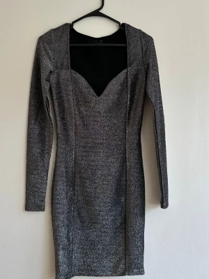 Glittrig silverfärgad bodyconklänning - Snygg silverglittrig bodyconklänning med långa ärmar och markerad sweetheart-urringning. Klänningen är figurnära och har en cool metallic look som verkligen sticker ut. Perfekt för dig som vill ha något extra till nästa utekväll. Materialet är stretchigt och följsamt.