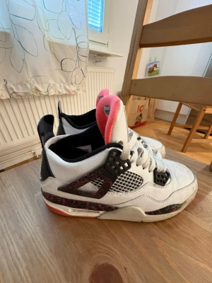 Air Jordan 4 Retro Vit/Svart/Rosa - Säljer ett par Air Jordan 4 Retro sneakers i vitt läder med svarta och rosa detaljer. Skorna har meshpaneler på sidorna, svart plastdetalj vid snörningen och rosa insida på plösen. Klassisk Jumpman-logga på plösen och robust sula med rosa inslag.