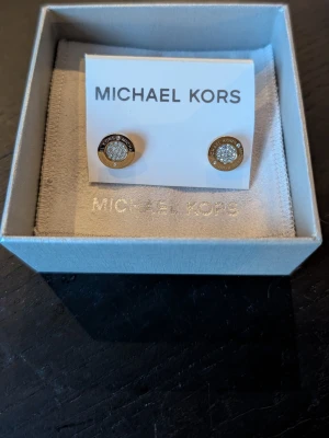 Guldiga örhängen Michael Kors - Snygga runda örhängen från Michael Kors i guldtonad metall med glittrande stenar i mitten. Klassisk design med logotyp runt kanten och gnistrande detaljer som ger en lyxig känsla. Perfekta för dig som gillar stilrena accessoarer med lite extra bling.