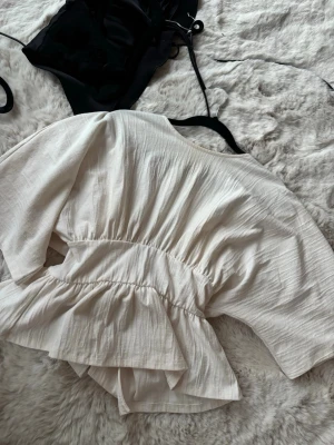 Gina topp - Snygg vit topp med breda knytband i ryggen. Blusen har en avslappnad passform och är gjord i ett mjukt, strukturerat tyg som ger en luftig känsla. Perfekt att styla med jeans eller kjol för en trendig look.