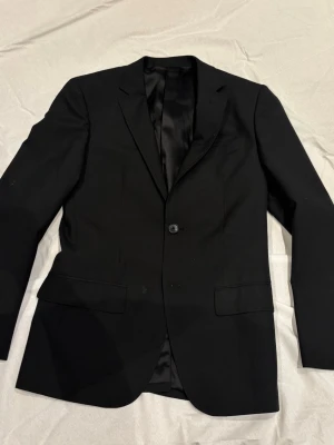 Klassisk svart kavaj med enkelknäppning - Stilren svart kavaj med enkelknäppning och två fickor framtill. Kavajen har smal passform, klassisk krage och helfodrad insida. Perfekt för en dressad look och passar till många olika stilar.