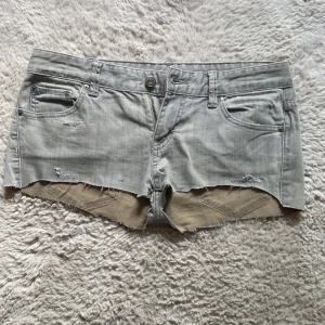 Grå jeansshorts från Armani Exchange - Snygga grå jeansshorts från Armani Exchange med råa kanter och slitna detaljer. Klassisk femficksmodell med låg midja och knappgylf. Perfekta för sommaren när du vill ha en chill och trendig look. Nypris 200kr