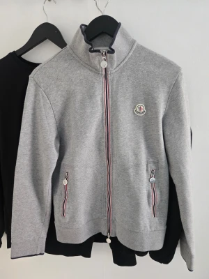 Grå zip hoodie från Moncler - Snygg grå zip hoodie från Moncler med hög krage och ikonisk Moncler-logga på bröstet. Två fickor med dragkedja och röd-vita detaljer. Tillverkad i mjukt sweatshirtmaterial, perfekt för en chill och stilren look.
