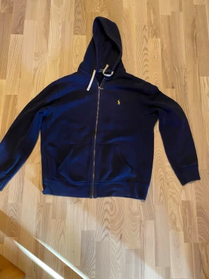 Mörkblå hoodie från Polo Ralph Lauren - Snygg mörkblå hoodie med dragkedja från Polo Ralph Lauren. Klassisk design med huva och vita dragsnören, samt den ikoniska gula loggan broderad på bröstet. Två fickor framtill och mjukt bomullsmaterial gör den både bekväm och stilren.