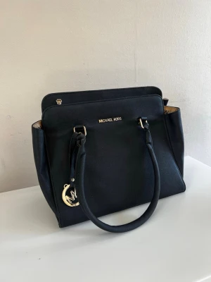 Svart handväska från Michael Kors  - Stilren svart handväska från Michael Kors (ej äkta) med guldfärgade detaljer och logotyp framtill. Väskan har två långa handtag, strukturerad form och en dekorativ MK-berlock. Insidan är fodrad i beige och väskan är tillverkad i tåligt konstläder. Sömmarna är lite slitna men annars i väldigt fint skick. 