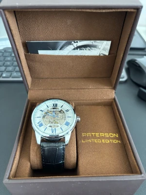 Paterson Limited Edition automatklocka - Snygg automatisk klocka från Paterson Limited Edition med silverfärgat boett, genomskinlig urtavla som visar mekaniken och blå visare. Svart läderarmband med krokodilstruktur. Perfekt för dig som gillar stilrena detaljer och mekaniska urverk.