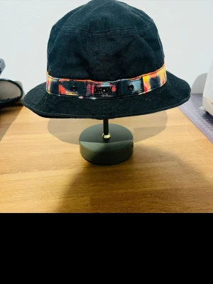 Svart bucket hat med färgglad detalj - Säljer en svart bucket hat från LIFUL med en unik färgglad banddetalj i orange, gul och rött runt hatten. Hatten är i mjukt tyg och har en avslappnad passform som ger en cool och trendig vibe. Perfekt accessoar för att lyfta din outfit.