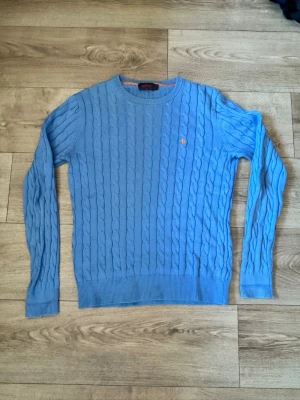Blå kabelstickad tröja från Morris  - Snygg blå kabelstickad tröja från Morris  med rund hals och ribbade muddar. Tröjan har ett orange broderat emblem på bröstet och är tillverkad i mjukt material. Perfekt för dig som gillar klassisk stil med en twist.