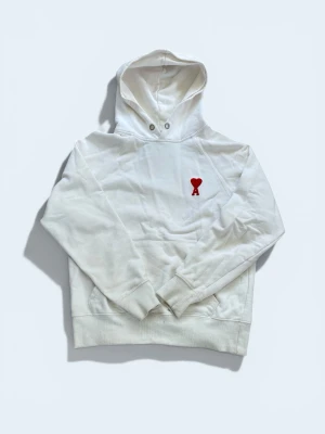 Vit hoodie  - Säljer en stilren vit hoodie från Ami Paris med deras ikoniska röda hjärta och A broderat på bröstet. Hoodien har huva med dragsko, känguruficka och ribbade muddar. Perfekt för en clean och trendig look.
