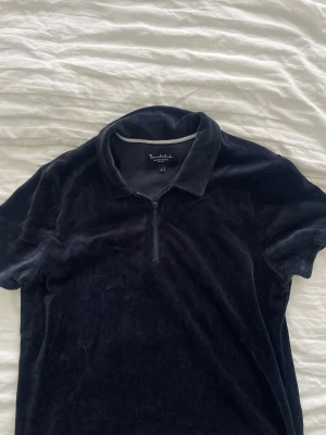 Mörkblå  polotopp i sammet med half zip - Mjuk och stilren svart polotopp från bondelid i sammet. Toppen har korta ärmar, klassisk krage och en half zip-dragkedja framtill för en modern touch. Perfekt för dig som gillar en avslappnad men trendig look.