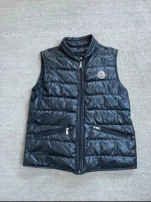 Moncler Väst - Säljer nu denna snygga Moncler väst! Den är i en glansig svart färg och i storlek 152 cm (12 år). Den är i mycket bra skick och självklart äkta! Nypris på västen är 4499 kr❌🤯Mitt pris är 1199 kr✅. Om du har frågor eller funderingar, eller om du vill ha fler bilder är det bara att fråga!🤗🤝