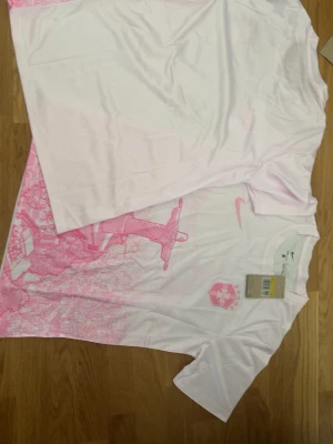 Vit och rosa Nike t-shirt - Säljer en vit t-shirt från Nike med rosa grafiskt tryck framtill och klassisk Nike-logga på bröstet. T-shirten har rund halsringning och korta ärmar. Perfekt för dig som gillar sportig och trendig stil.