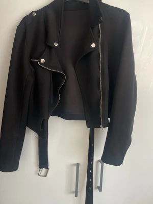 Svart biker skinnjacka med bälte - Snygg svart skinnjacka i klassisk bikerstil med silvriga dragkedjor och knappar. Jackan har ett avtagbart bälte i midjan och dragkedjor vid ärmsluten för extra edge. Kortare modell som passar perfekt till jeans eller klänning.