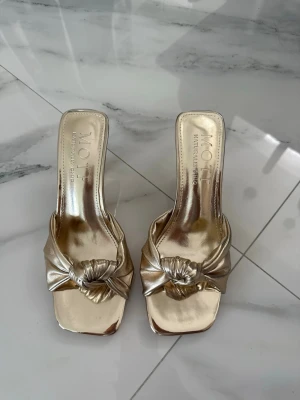 Guldiga knutpumps  - Säljer ett par trendiga guldiga pumps med öppen tå och fyrkantig framdel. Skorna har en snygg knutdetalj framtill och en smal klack. Tillverkade i ett glansigt material som ger en lyxig känsla. Perfekta för dig som vill sticka ut.