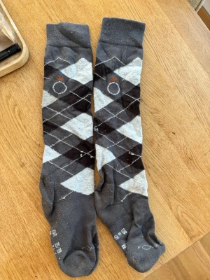 Grå rutig strumpor från Rouch - Snygga knälånga strumpor i grått med klassiskt rutig argyle-mönster i svart, vitt och ljusgrått. Tillverkade i mjuk bomullsmix som sitter skönt på foten. Perfekta för dig som vill ha en stilren men ändå lite rolig detalj till outfiten.