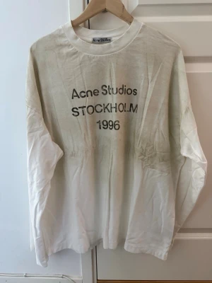 Vit långärmad Acne Studios t-shirt - Vit långärmad t-shirt från Acne Studios med tryck på bröstet där det står 'Acne Studios STOCKHOLM 1996'. Klassisk rund hals och relaxed fit. Perfekt för dig som gillar minimalistisk och stilren design.