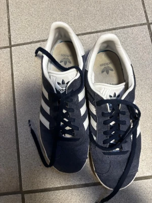 Marinblå Adidas sneakers med vita ränder - Säljer ett par klassiska marinblå Adidas sneakers med de ikoniska tre vita ränderna på sidan. Skorna har vit sula och vit häl, samt mörkblå snören. Ovandelen är i mockaliknande material och insidan är vit med Adidas-logga.