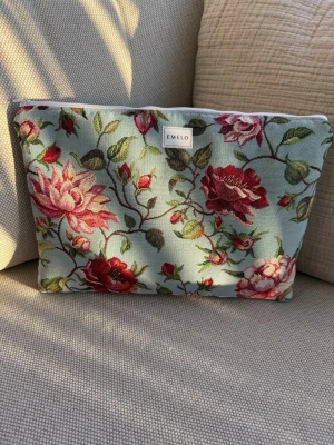 Blommig datorväska från Emelo - Snygg datorväska från Emelo i tyg med blommigt mönster i rött, rosa och grönt på ljusblå botten. Väskan har dragkedja upptill och en mjuk, rektangulär form. Perfekt för dig som vill ha en unik och färgglad accessoar till din laptop. Passar laptop med maxmått: 32x24 cm