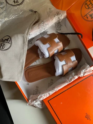 Bruna sandaler med H-design - Säljer ett par trendiga bruna sandaler med öppen tå och bred rem i H-form. Sandalerna har vita kontrastsömmar och är tillverkade i slätt konstläder. Perfekta för en avslappnad och stilren look.
