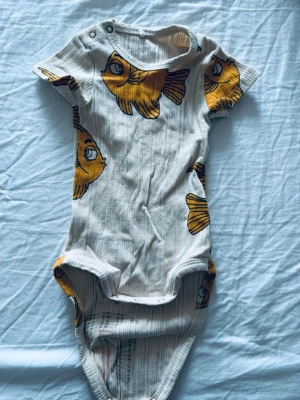Vit body med fiskar från Mini Rodini - Supersöt vit ribbad body från Mini Rodini med korta ärmar och tryckknappar vid axeln. Bodyn har ett coolt mönster med stora gula fiskar och är gjord i mjukt, stretchigt material. Perfekt för dig som gillar lekfull design och sköna plagg.
