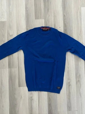 Blå tröja från Superdry - Snygg blå tröja från Superdry i bomull. Tröjan har rund halsringning, långa ärmar och ribbade muddar vid ärmslut och nederkant. En liten Superdry-logga sitter nere vid fållen. Perfekt för dig som gillar stilren och enkel design.