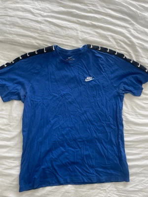 Blå Nike t-shirt med logga - Snygg blå t-shirt från Nike med vit logga på bröstet och svarta band med vita swoosh-detaljer längs axlarna. Klassisk passform och rund halsringning. 