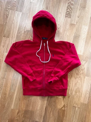 Röd hoodie från Polo Ralph Lauren - Tjena, jag säljer denna röda zip up från Ralph Lauren med vita snören. Jag köpte den från vinted i bra skick men den har aldrig använts eftersom den är för liten, men den har inga som helst fel och materialet är fortfarande mjukt.  Storlek S  Röd   Ralph lauren   Mått 📏  Längd 60cm  Axelbredd 49cm.       Hör av dig om du undrar något!