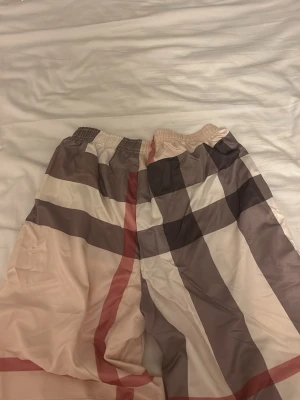 Rutig shorts med beige och bruna toner - Säljer ett par rutiga shorts med bred resår i midjan. Shortsen har ett klassiskt mönster i beige, brunt, vitt och röda detaljer. Perfekta för sommaren och har en avslappnad passform. Materialet känns lätt och luftigt.